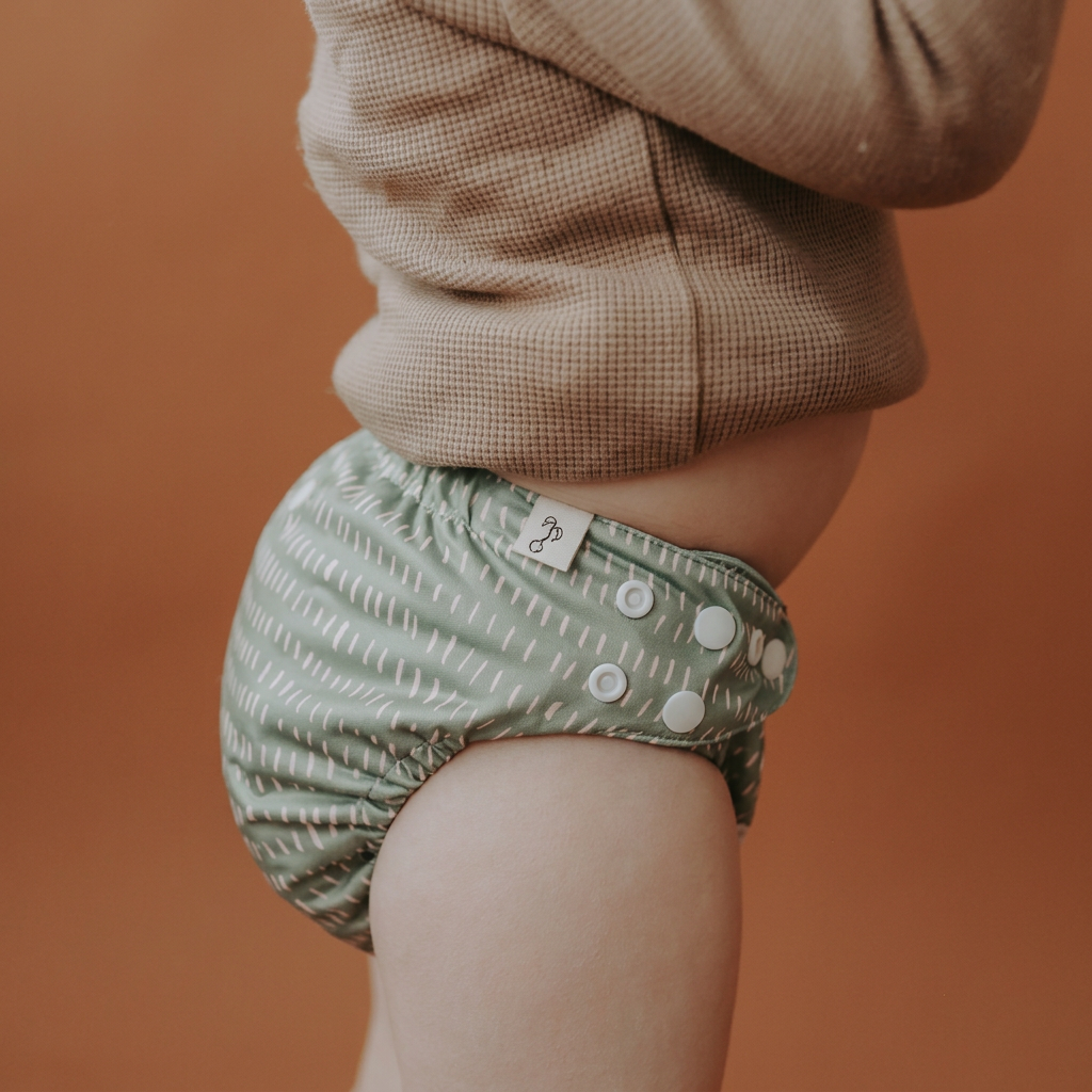 A close-up side view of a child wearing a sage green reusable cloth diaper with a white dotted pattern and white snap buttons, paired with a brown textured top against a solid warm brown background.