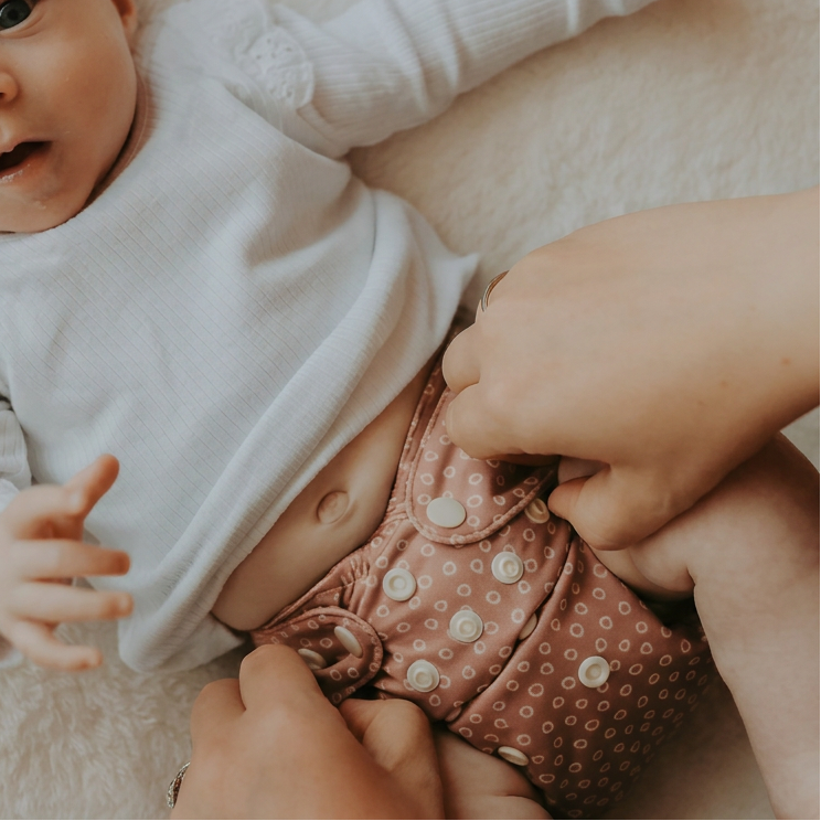 A baby lying on a soft cream blanket, wearing a white long-sleeve top and a dusty pink reusable cloth diaper with white polka dots and snap closures, while an adult's hands adjust the diaper.