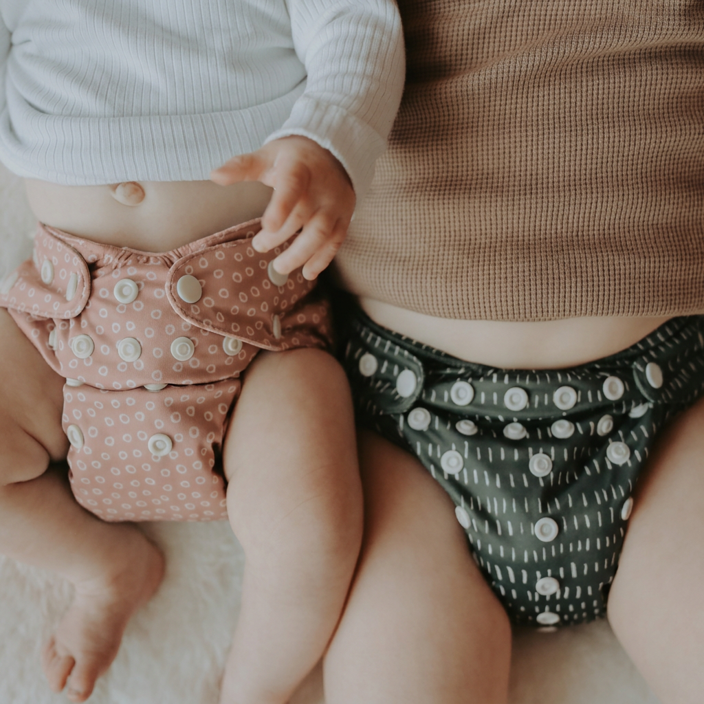 Two babies wearing reusable patterned cloth diapers, shown in a close-up lifestyle shot. One diaper is dusty pink with small white polka dots, and the other is dark gray with white vertical stripes and white dots.