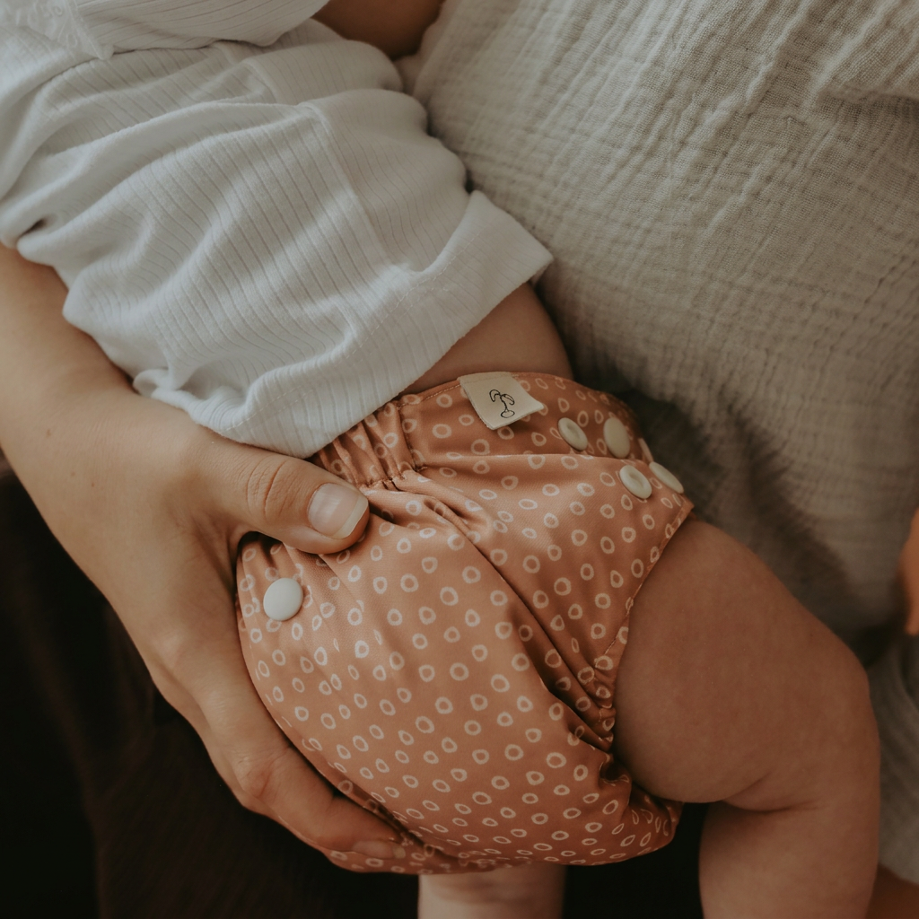 A close-up lifestyle shot of a baby wearing a dusty rose reusable cloth diaper with a white circle pattern and snap closures, held by an adult.