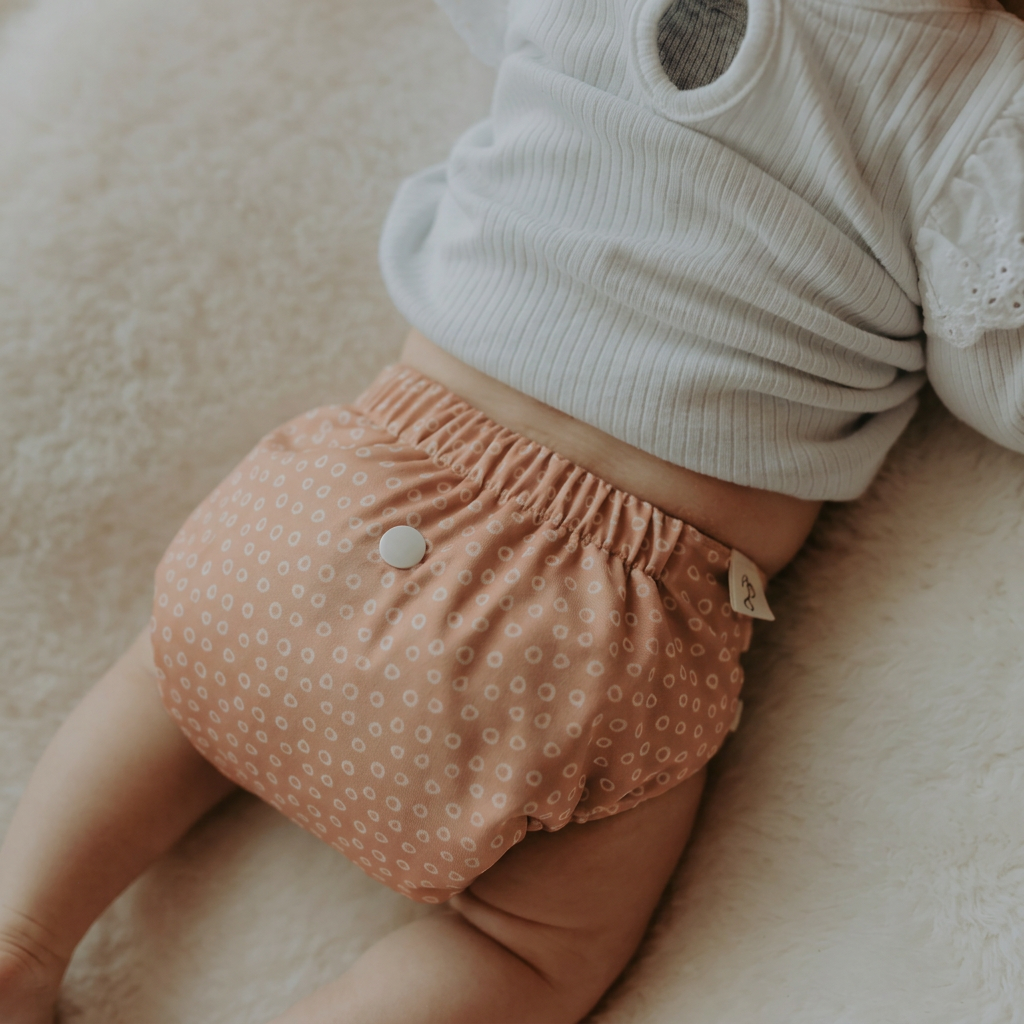 A close-up of a baby wearing dusty pink patterned cloth diaper cover with a snap button, paired with a white ribbed bodysuit, lying on a soft cream-colored blanket.